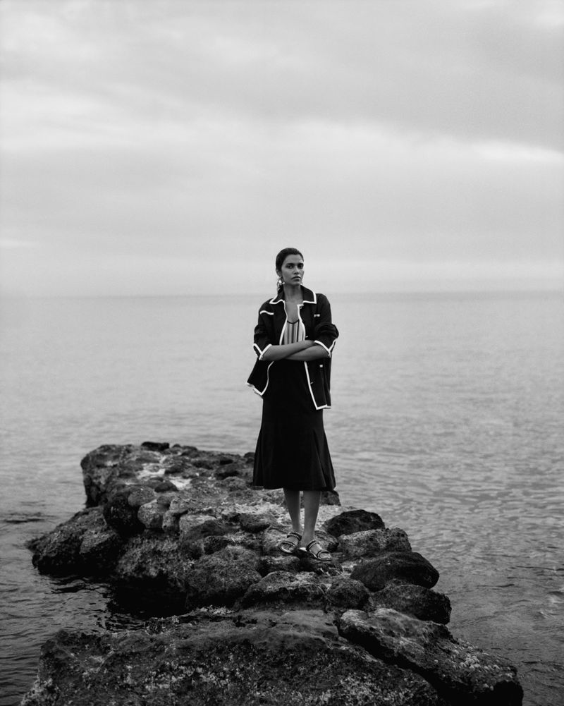 Model Pooja Mor standing on rocky coastal rocks by the ocean in a black jacket and skirt for Grazia UK Shore Things editorial.