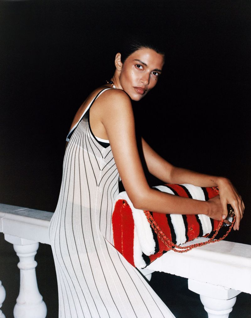 A woman in a striped dress leans on a railing, holding a striped bag against a dark background in a fashion-style photograph.