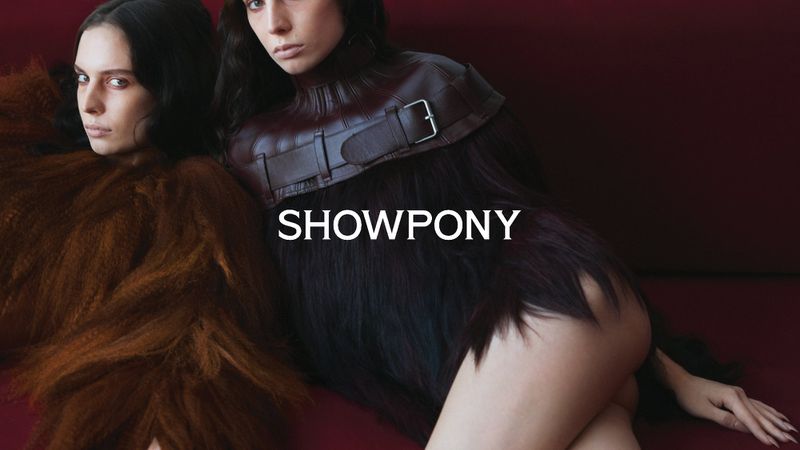 Two women pose indoors in a studio setting, one in a brown fur coat and the other in a black fur and leather harness-like top.