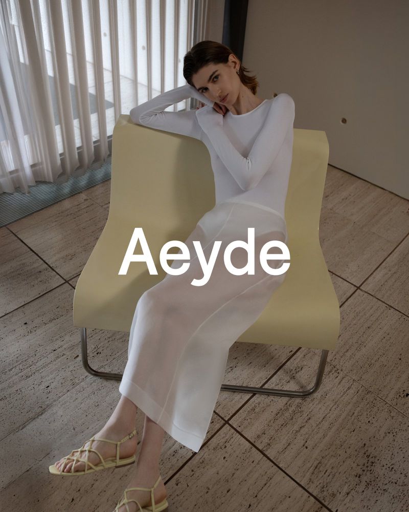 A fair-skinned woman is sitting in a pale yellow chair, wearing a white, long-sleeved shirt, a white skirt, and yellow strappy sandals.