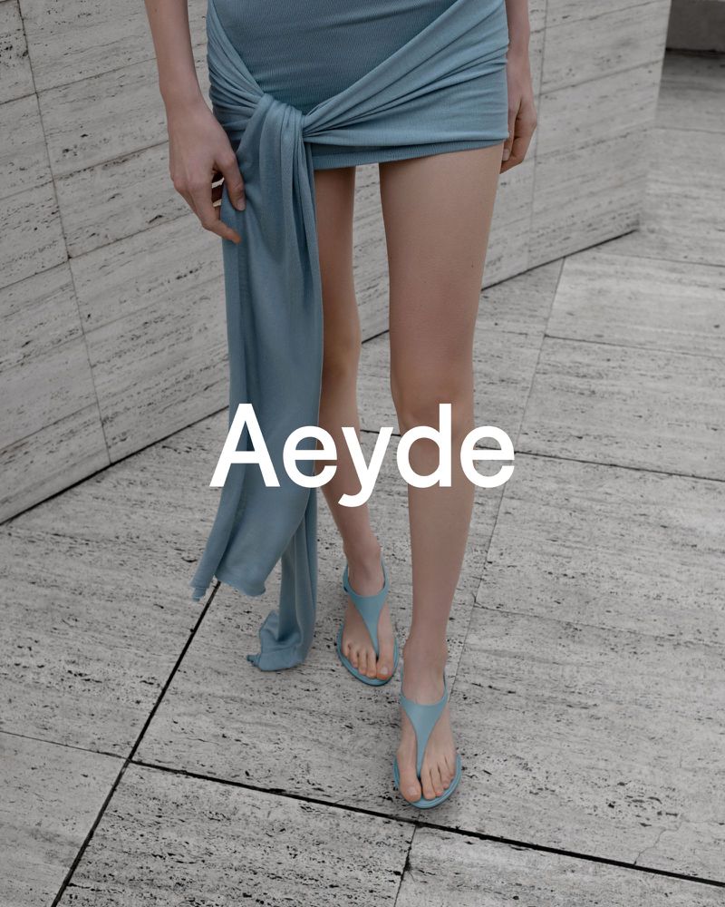 Model wearing blue dress and thong sandals standing outdoors on stone tile ground for Aeyde