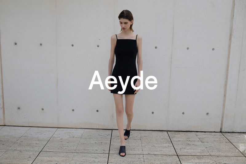 Model walking forward wearing a black slip dress and mules against a concrete wall for Aeyde High Summer 26