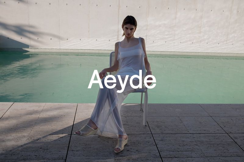 Model wearing a sheer white dress seated on a chair by a swimming pool for Aeyde High Summer 26 campaign