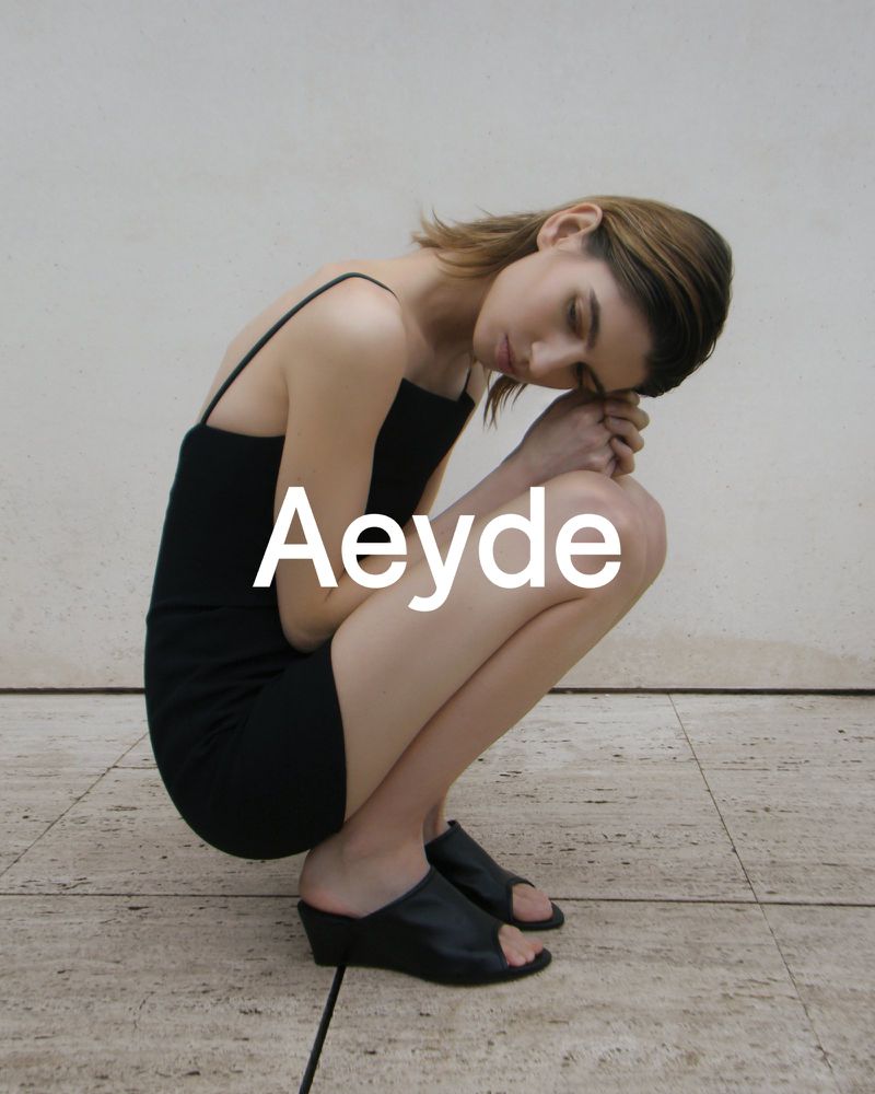 Model in a black slip dress and black mule sandals crouching against a neutral stone background for Aeyde