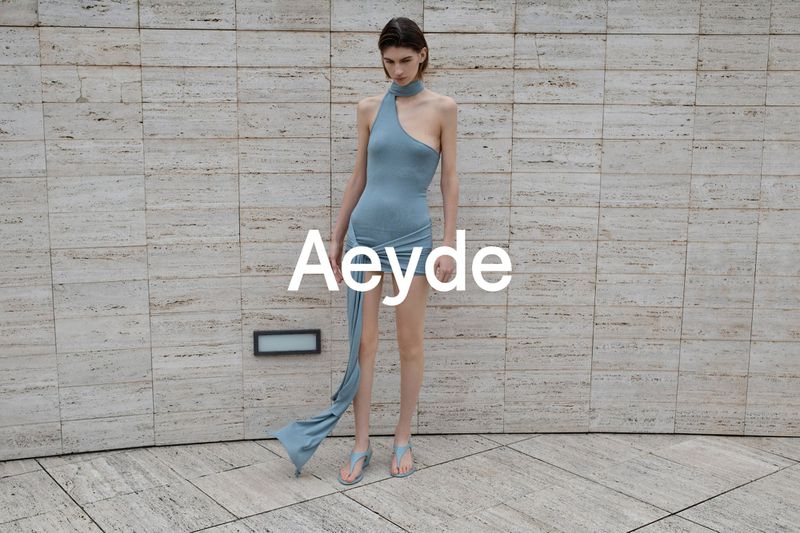 Model in a blue asymmetric dress and sandals standing against a tiled stone wall for Aeyde