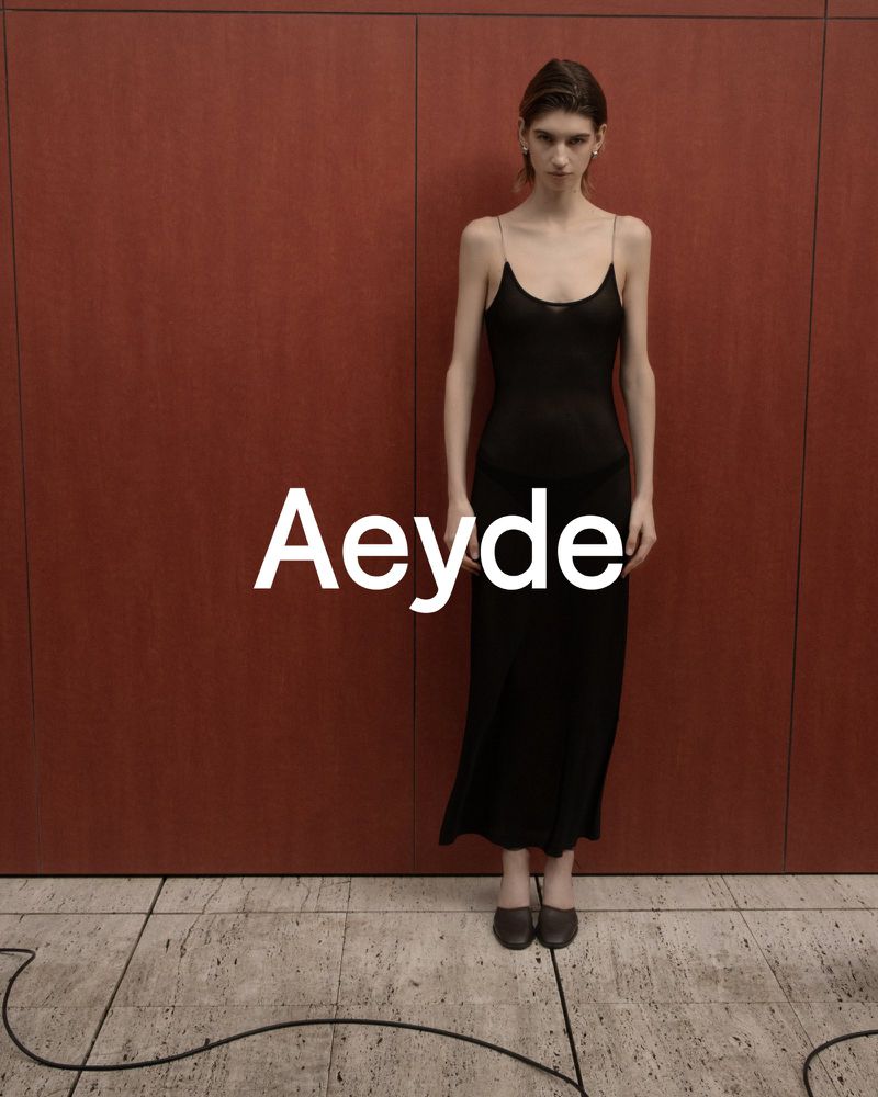Model standing against a reddish-brown paneled wall wearing a black slip dress and dark shoes for Aeyde.
