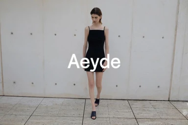 Model walking forward wearing a black slip dress and mules against a concrete wall for Aeyde High Summer 26