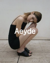 Model in a black slip dress and black mule sandals crouching against a neutral stone background for Aeyde