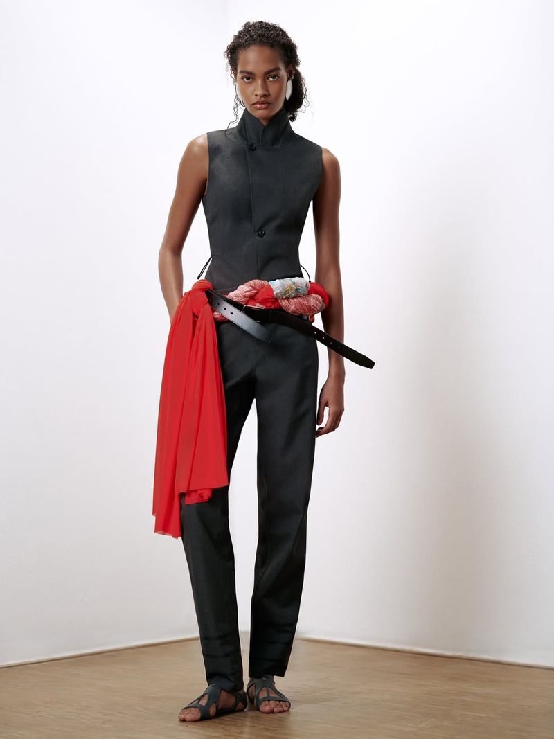Anok Yai wearing a dark sleeveless top, trousers, and a red sash against a neutral backdrop for Coperni.