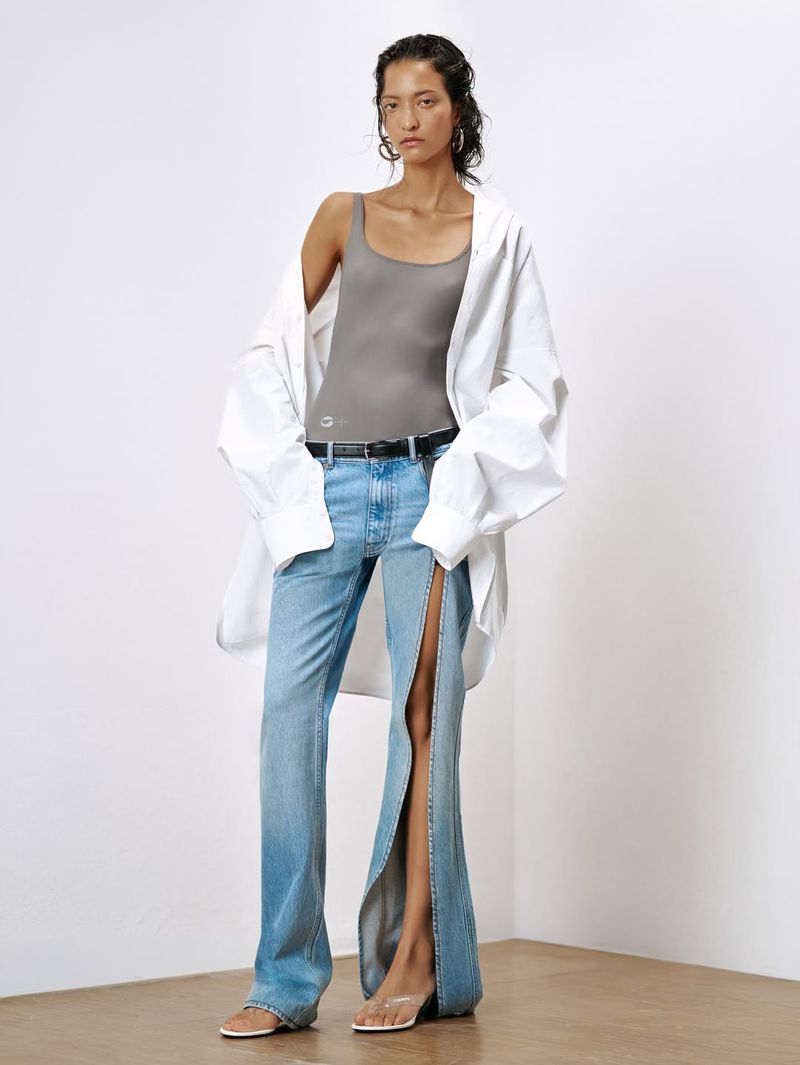 Model standing against a white studio wall wearing a grey bodysuit, unbuttoned white shirt, and slit blue jeans.