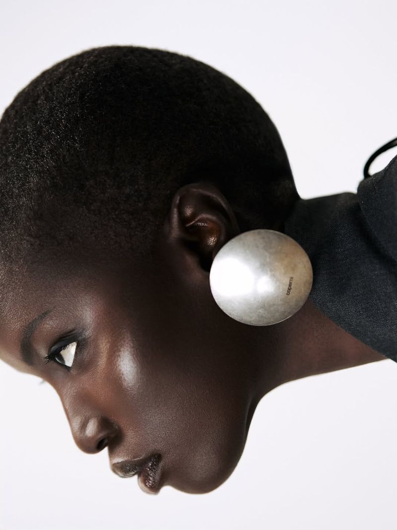 Close-up profile of model Anok Yai wearing a large silver earring against a white studio background