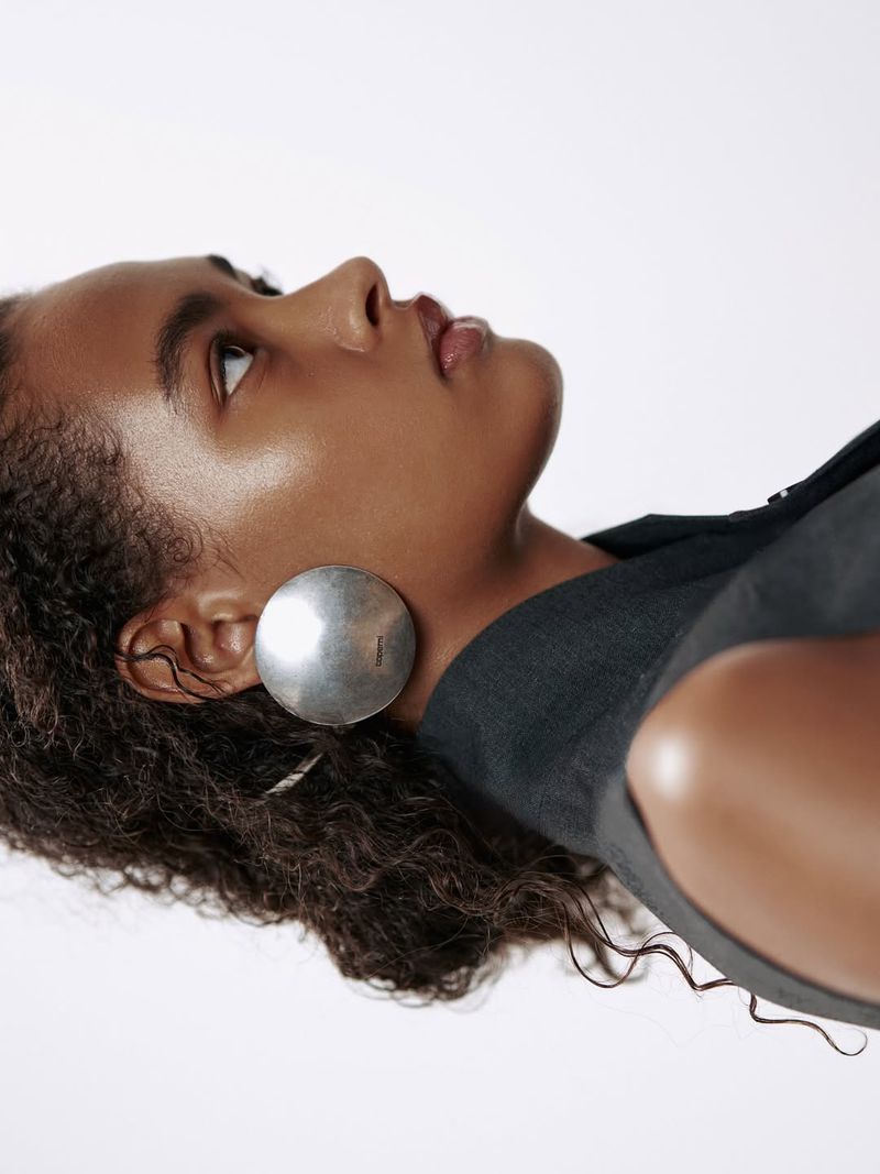 Anok Yai wearing large round silver earrings and a dark sleeveless top against a clean white background