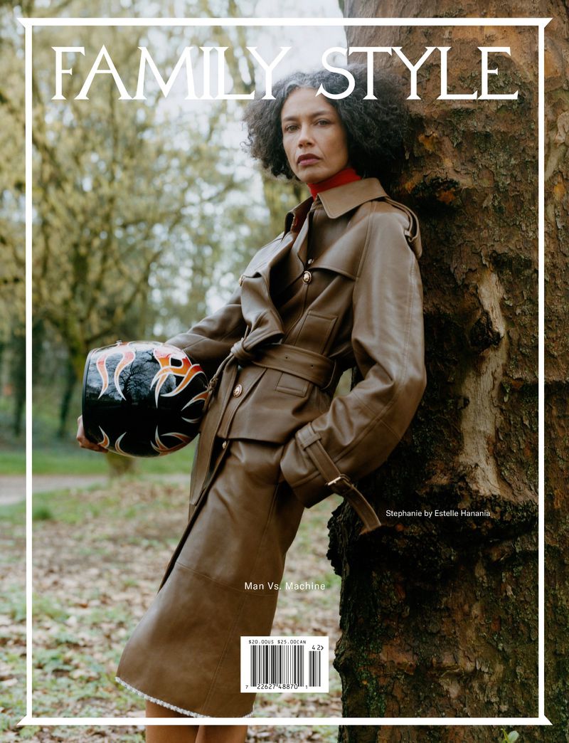Woman leaning against a tree trunk wearing a brown leather coat and holding a patterned helmet