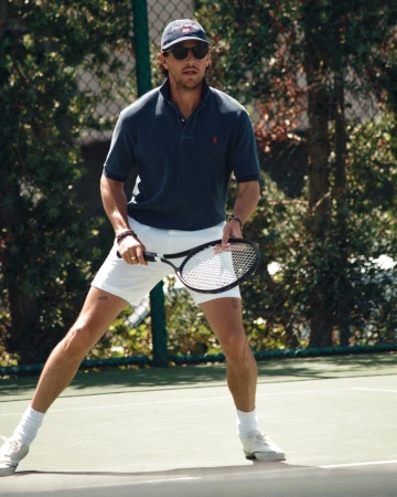 Man playing tennis wearing sunglasses, cap, polo shirt and white shorts holding a racket on a court
