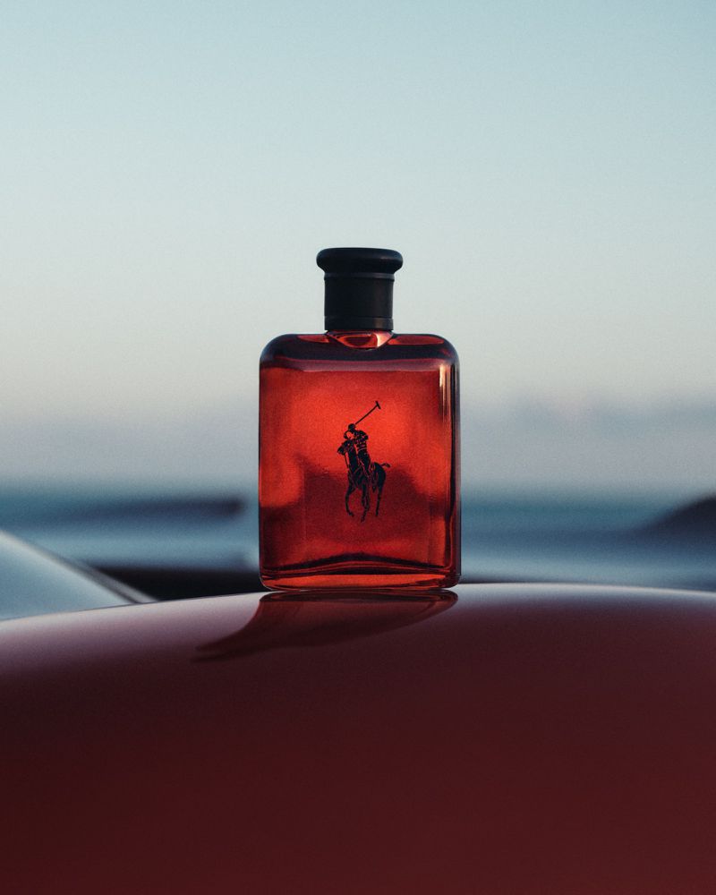 Red perfume bottle with dark cap resting on a glossy red car surface against an outdoor horizon background
