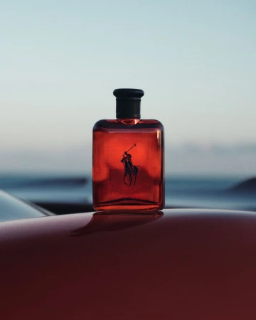 Red perfume bottle with dark cap resting on a glossy red car surface against an outdoor horizon background