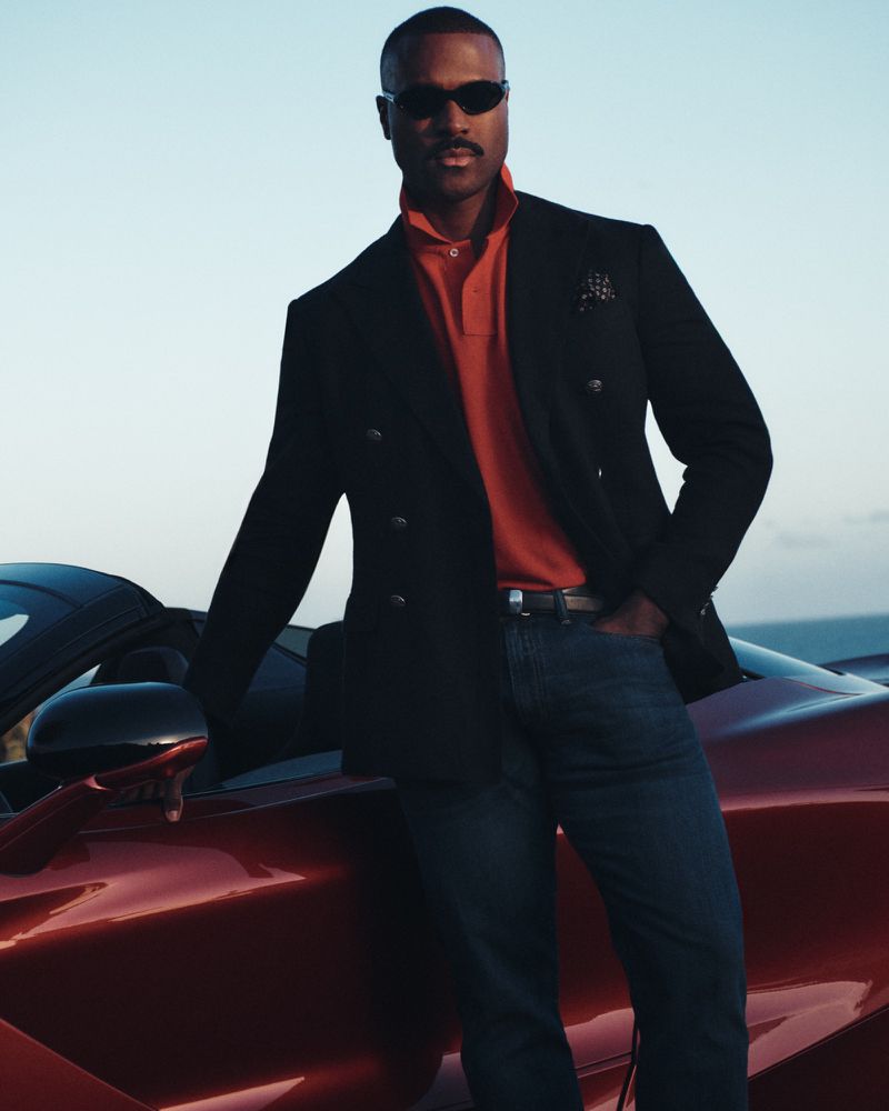 Man wearing sunglasses and a black blazer leaning against a red sports car outdoors at sunset