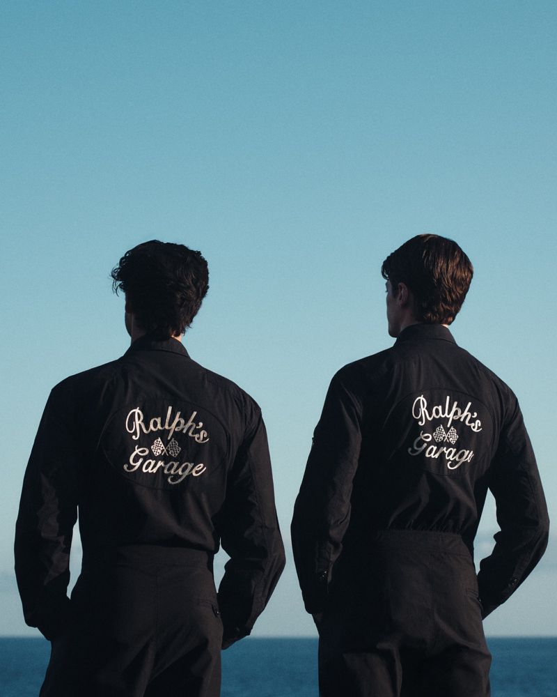 Two men from behind wearing black shirts with embroidered text against an ocean and clear sky background