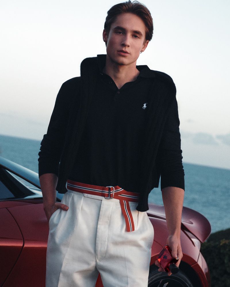 Young man in a black polo shirt and white trousers leaning against a red car with the ocean behind him.