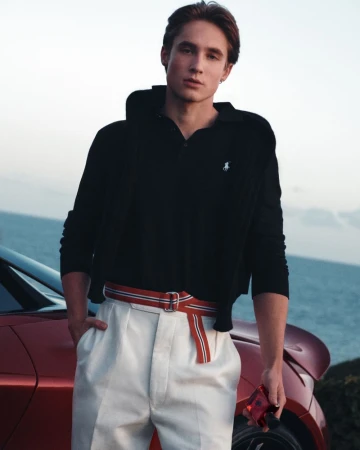 Young man in a black polo shirt and white trousers leaning against a red car with the ocean behind him.