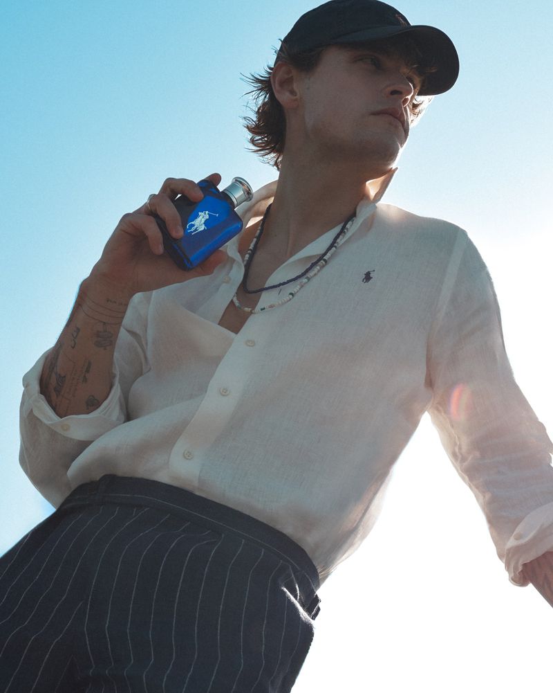 Young man in a white linen shirt and black cap holding a blue fragrance bottle against a bright sky