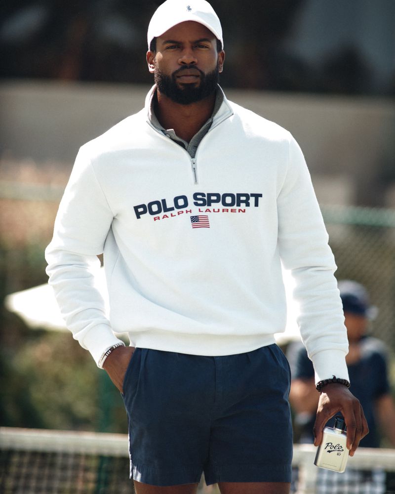 Black man wearing a white quarter-zip sweatshirt and holding a Ralph Lauren Polo Sport fragrance bottle outdoors.
