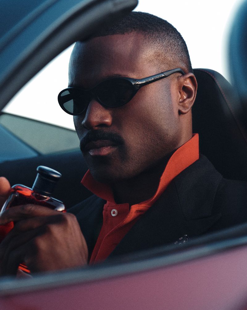 Black man wearing sunglasses and a jacket holding a Ralph Lauren fragrance bottle inside a car