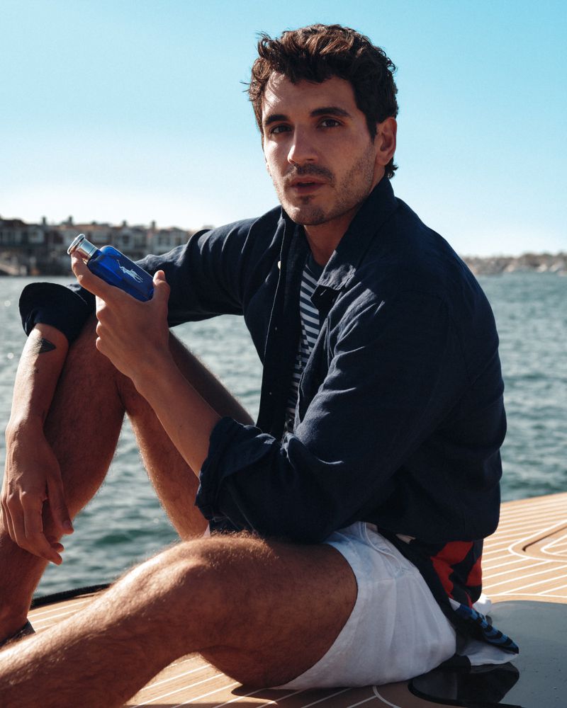 Man holding a blue perfume bottle while seated on a wooden dock by the water under a clear sky.