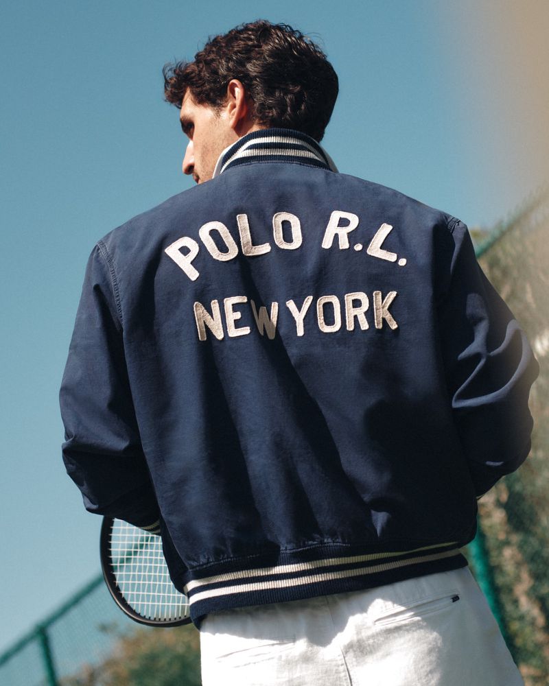 A man wearing a blue varsity jacket with Polo RL New York lettering holding a tennis racket outdoors against a clear blue sky