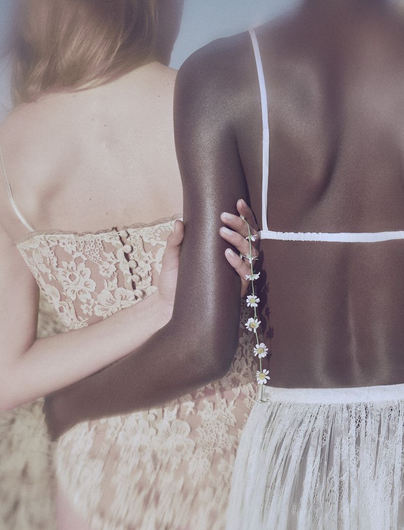 Two women embracing from behind wearing white lace garments, one holding a delicate chain of small white flowers.