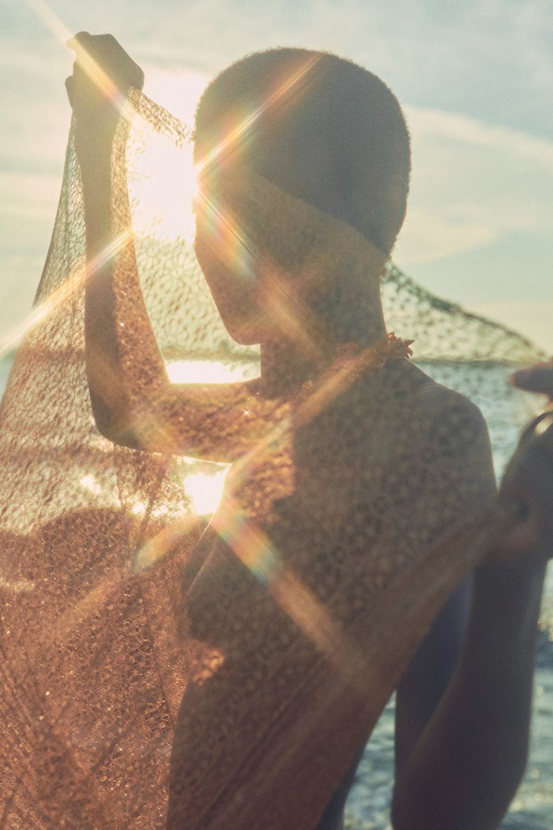Model holding a textured fabric against a bright sunset sky by the water for Ulla Johnson ULLA eau de parfum.