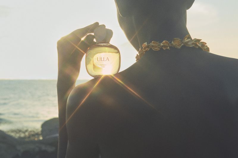 Person holding up a glass perfume bottle against a glowing sunset over the ocean with lens flare.
