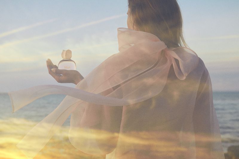 Woman in sheer pink organza blouse holding Ulla Johnson eau de parfum bottle against ocean background