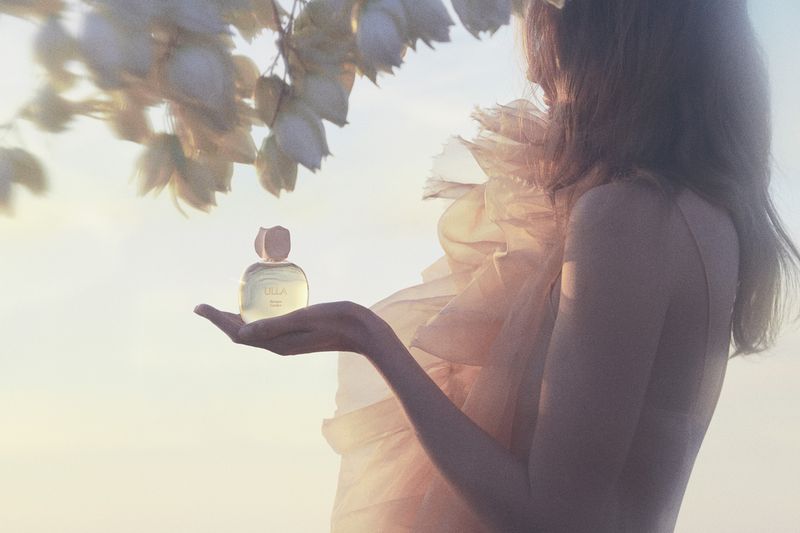 Woman in sheer ruffled dress holding perfume bottle under flowering branches at sunset for Ulla Johnson ULLA eau de parfum