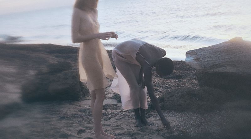 Two women in sheer slip dresses standing on a rocky sandy beach by the ocean under soft natural light.
