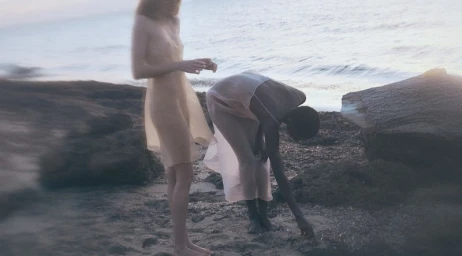Two women in sheer slip dresses standing on a rocky sandy beach by the ocean under soft natural light.