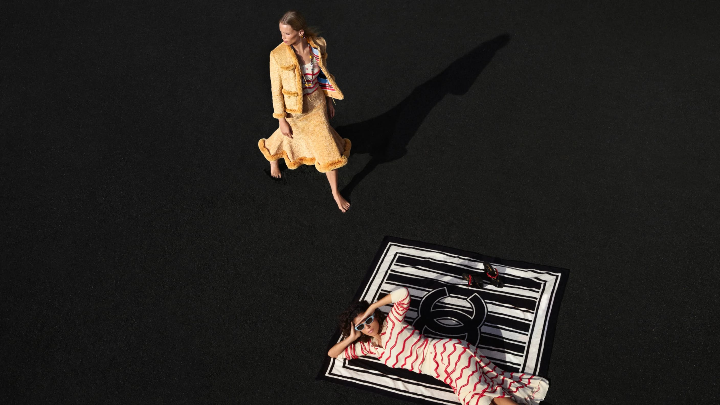Two models on a dark surface, one standing in yellow tweed apparel and another reclining on a black and white blanket.