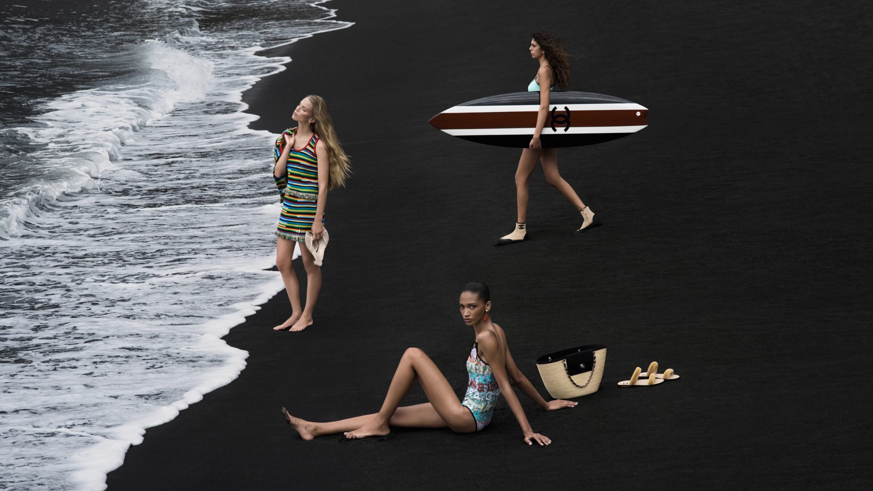 Three models on a dramatic black sand beach with ocean waves, carrying a Chanel surfboard and straw tote bag.