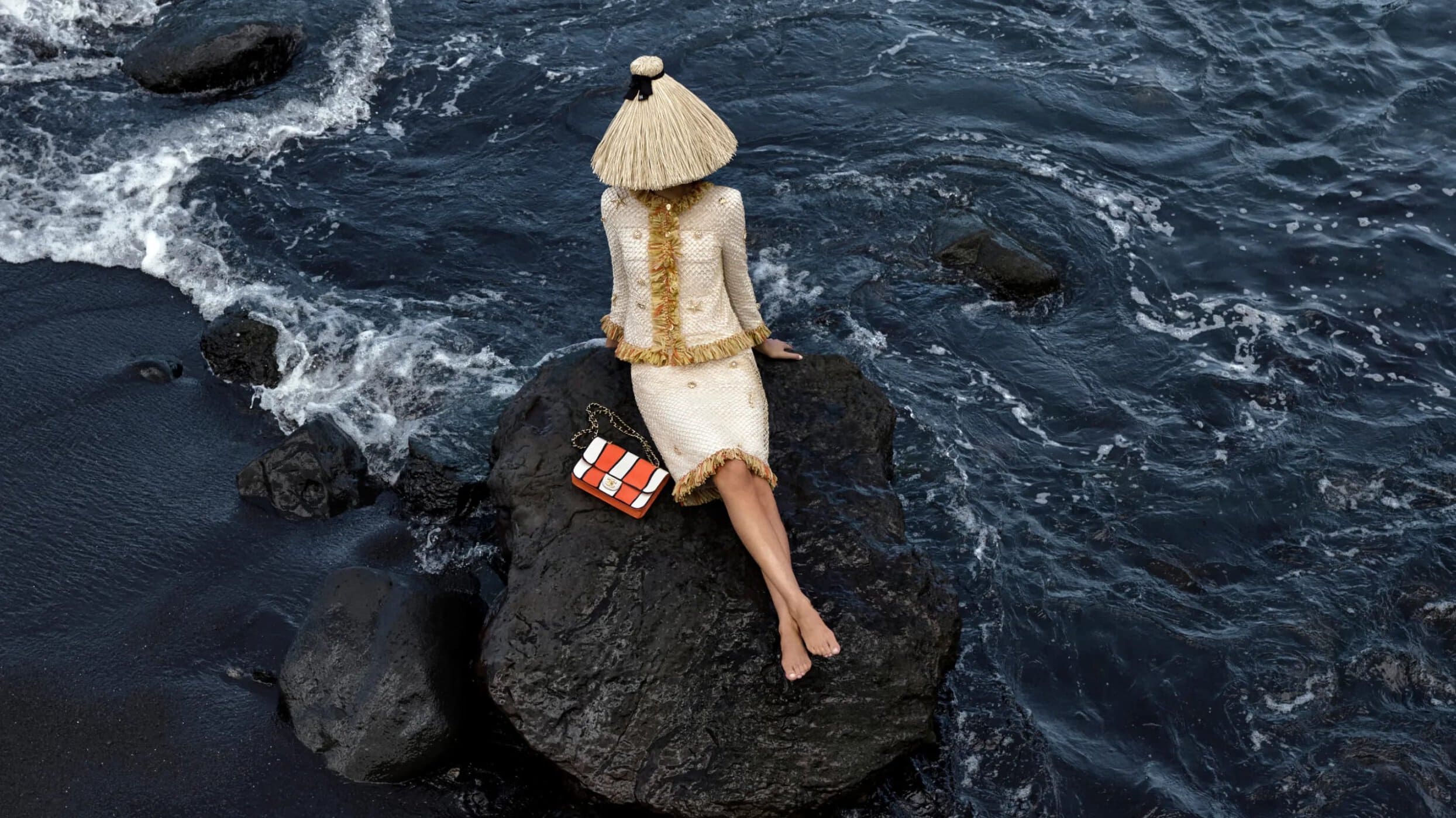 Adult female in a cream tweed skirt suit and matching sun hat sits on a rock at a black sand beach with a patterned handbag.