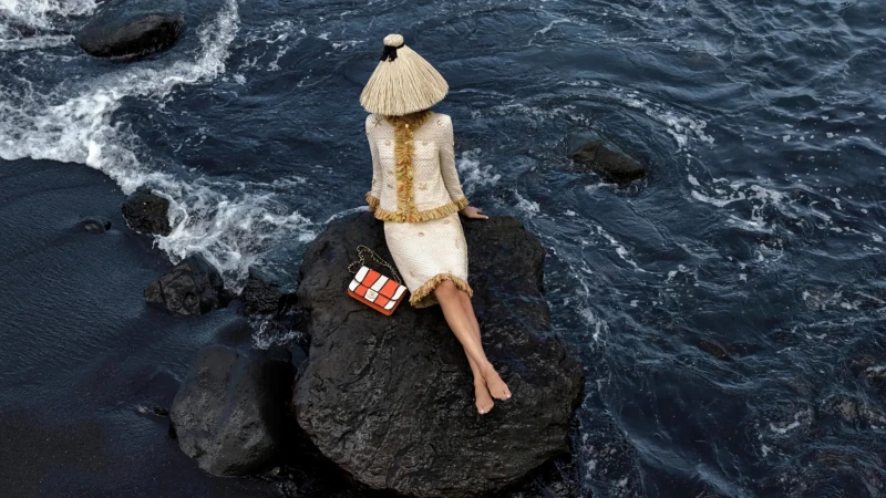 Adult female in a cream tweed skirt suit and matching sun hat sits on a rock at a black sand beach with a patterned handbag.