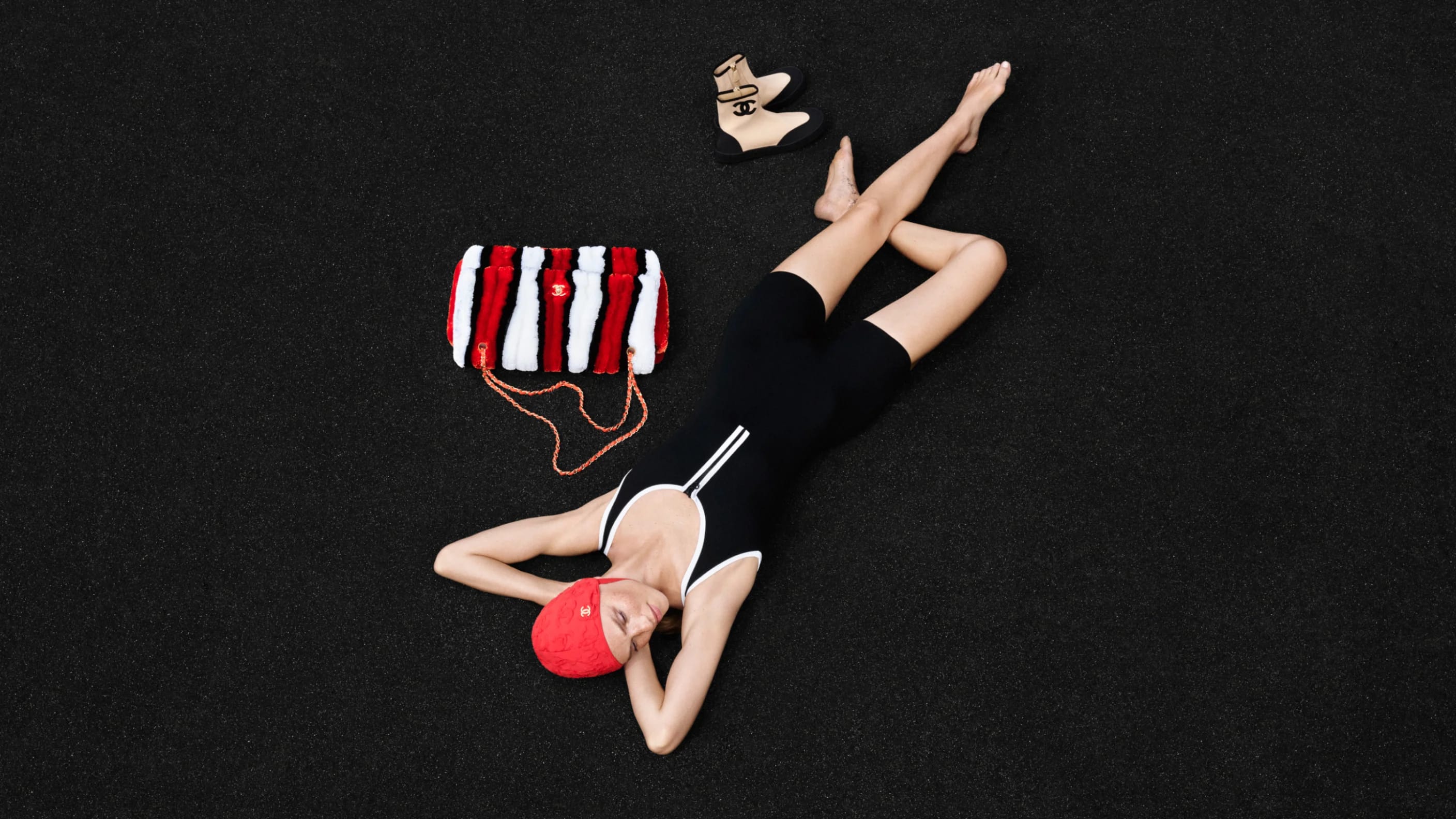 Model lying on dark background wearing black swimsuit and red swim cap beside striped fluffy handbag and boots