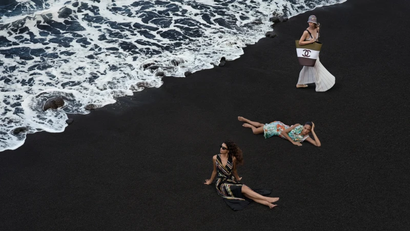 Three models on a dark volcanic sandy beach by crashing ocean waves, captured from a high-angle aerial perspective.