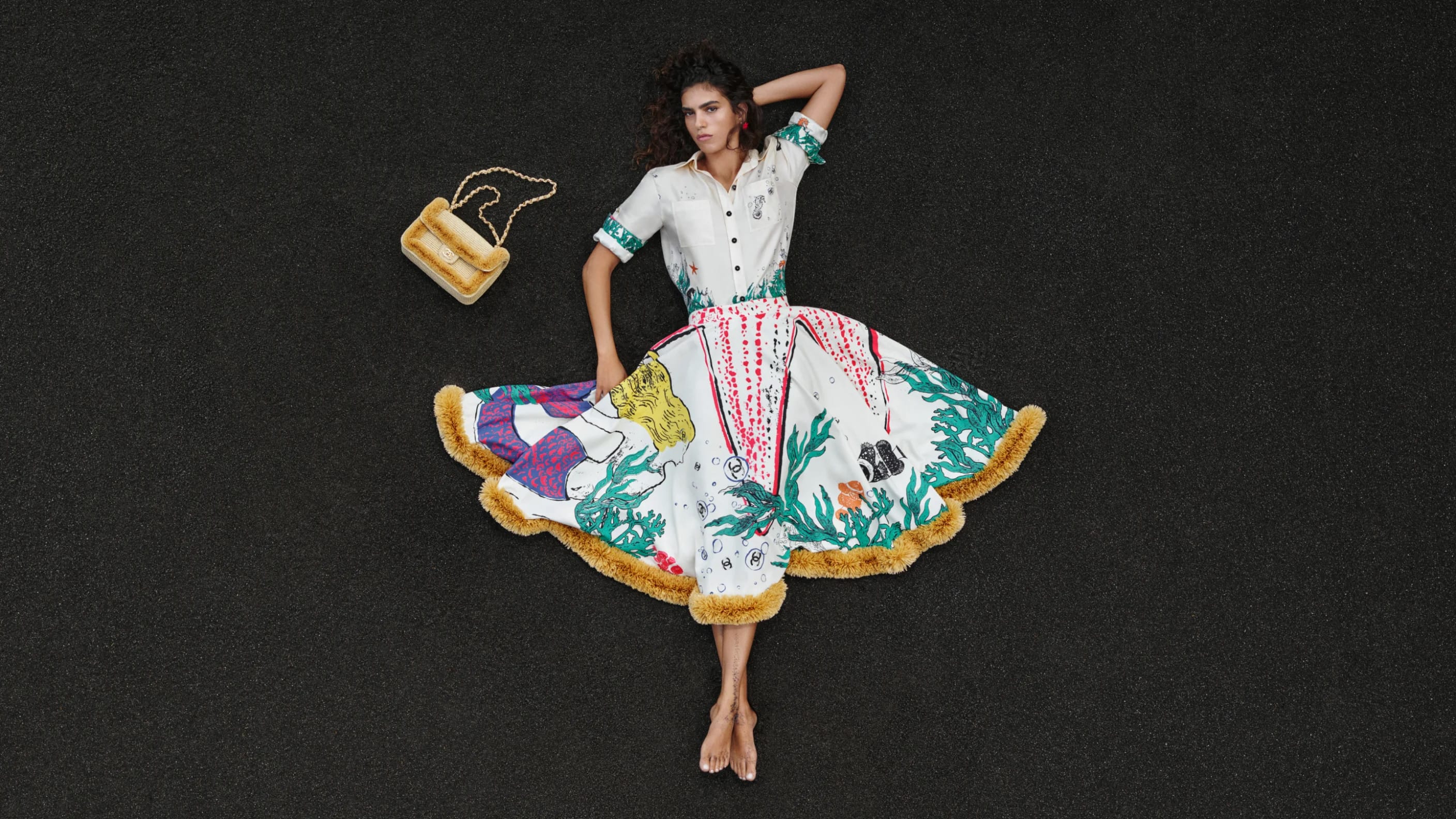 Model lying on dark textured asphalt wearing a white patterned shirt and full skirt with a matching handbag nearby