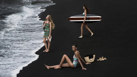 Three models on a dramatic black sand beach with ocean waves, carrying a Chanel surfboard and straw tote bag.