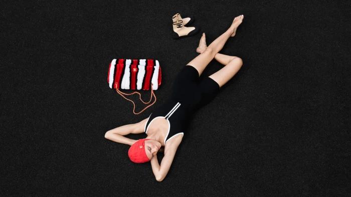 Model lying on dark background wearing black swimsuit and red swim cap beside striped fluffy handbag and boots