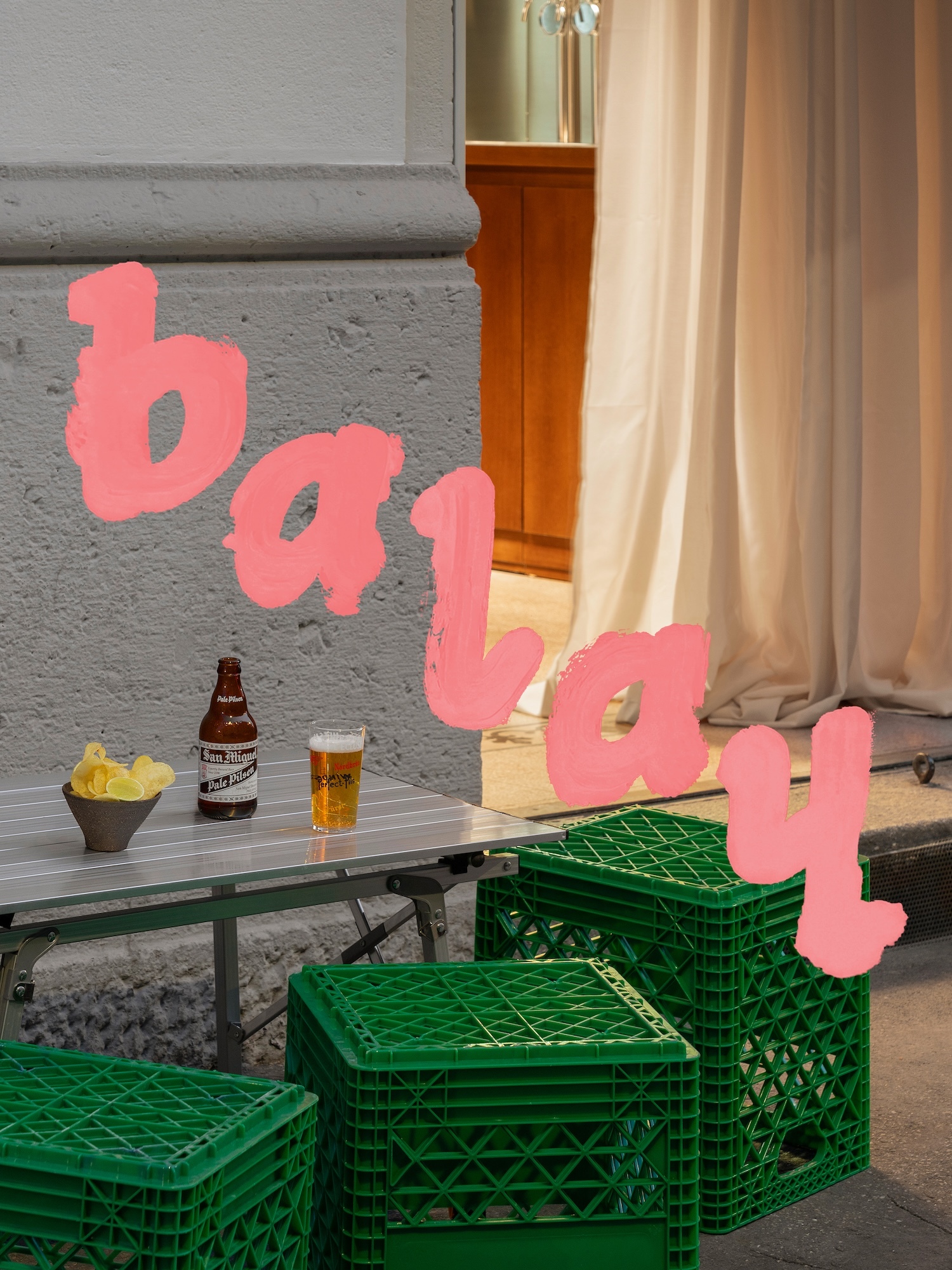 Balay brand identity image featuring a small outdoor table with a beer bottle and glass, and green plastic crates seating.