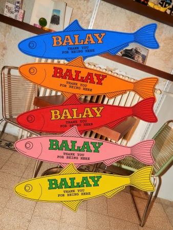 Five brightly colored fish-shaped wooden cutouts with Balay branding stacked vertically in an indoor setting.