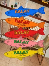 Five brightly colored fish-shaped wooden cutouts with Balay branding stacked vertically in an indoor setting.
