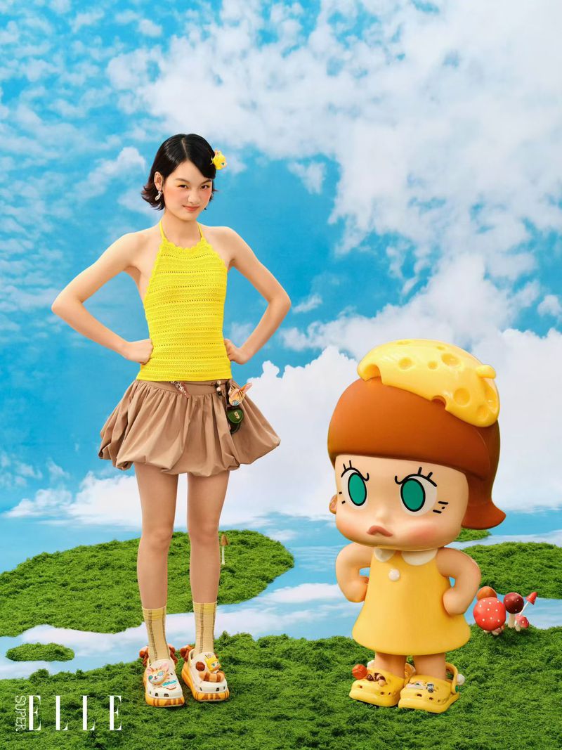 A woman in a yellow halter top and brown skirt stands beside a large cartoon figurine, both in an outdoor, grassy setting.