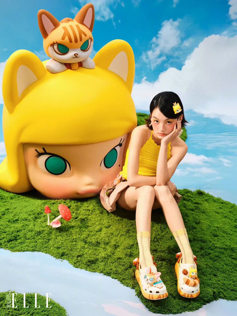 Woman in yellow top and patterned clogs sitting on moss next to a giant stylized yellow figure under a cloudy sky.